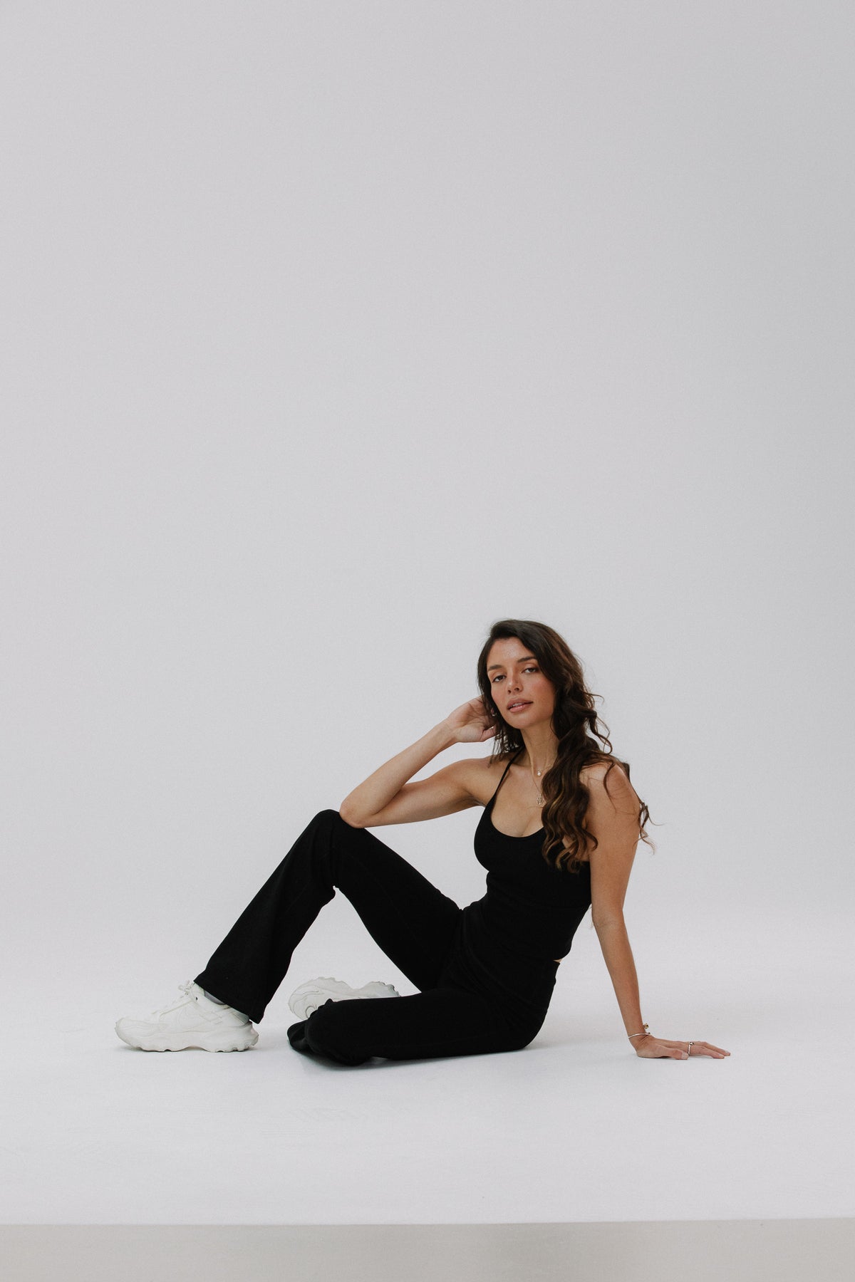 A woman with long brown hair sits on a white studio floor wearing a black cotton tank top and black cotton flared pants