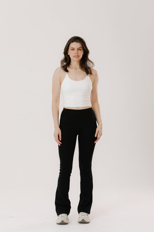 A tall woman stands is wearing a white cottonr tank top and tall length black flared pants, with white sneakers.