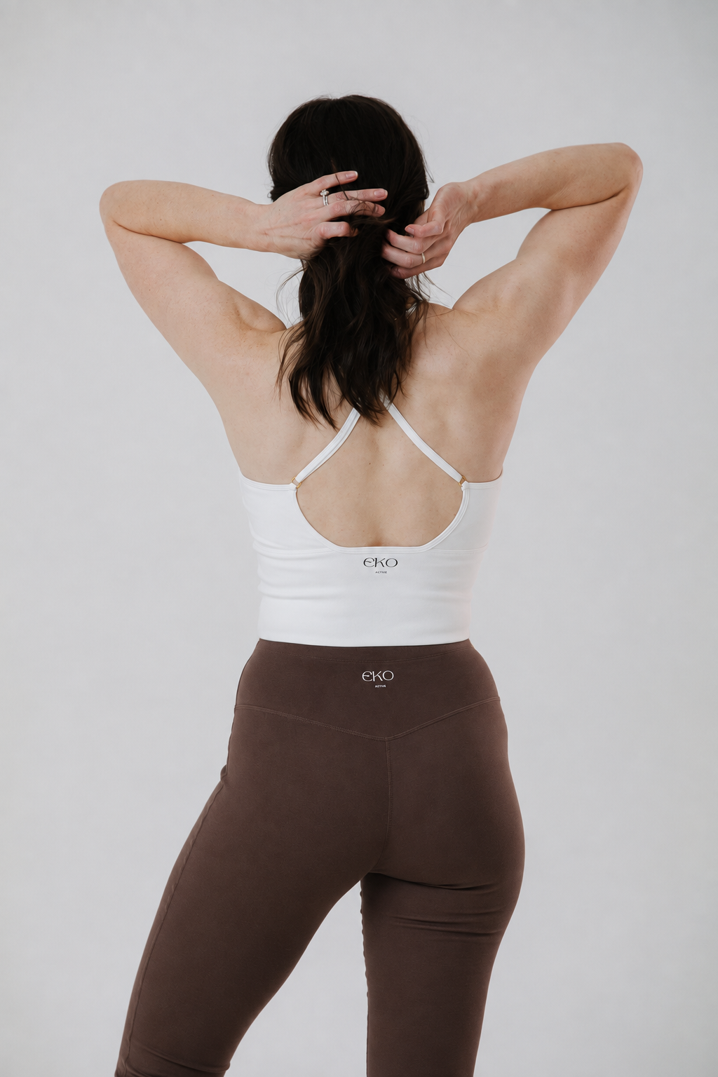 A woman shown from behind wearing a white sports bra with a low open-back design and brown leggings, standing against a light gray studio background.