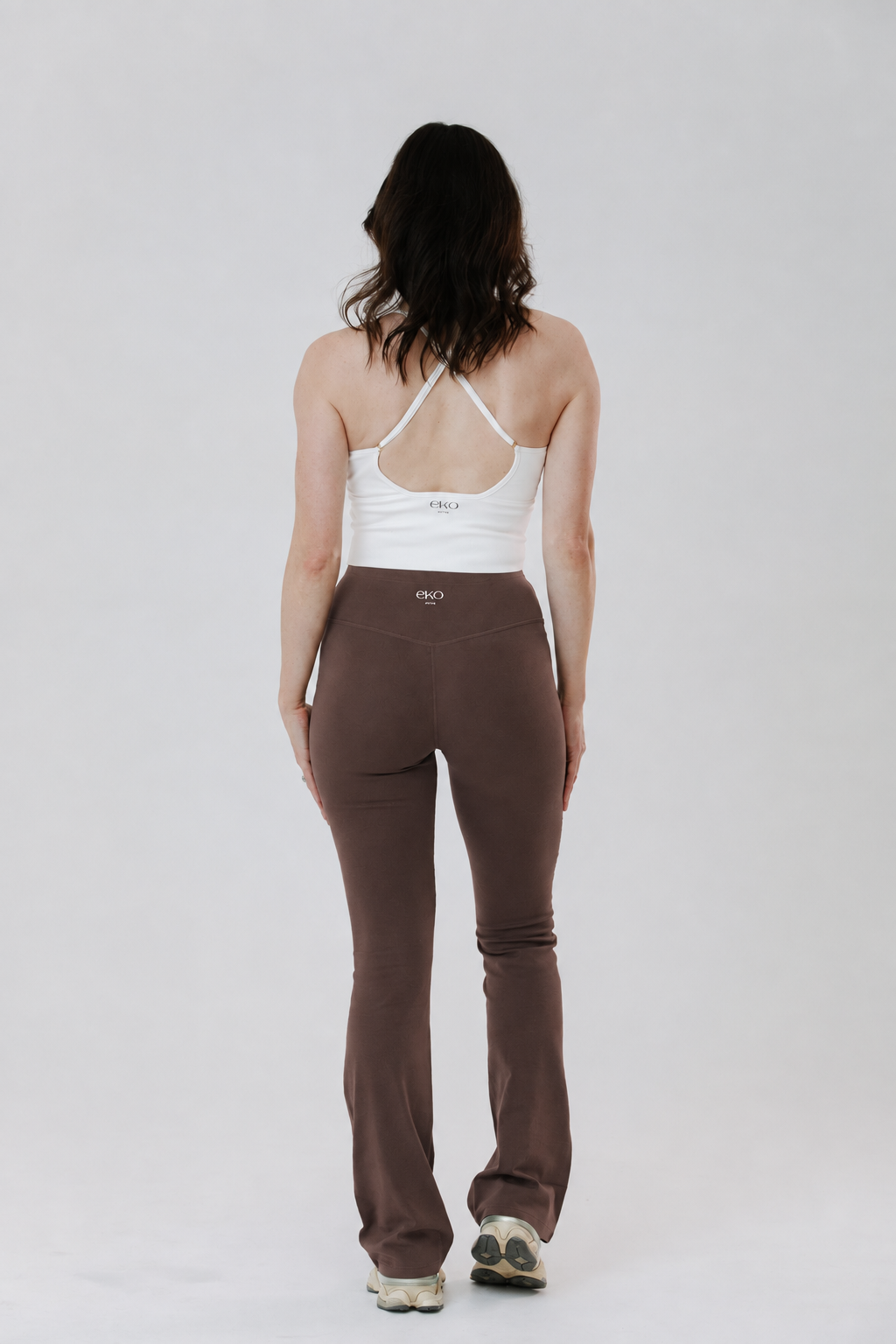 A tall woman viewed from behind wearing brown cotton high-waisted flared leggings and a white racerback tank top, standing against a light gray studio background.