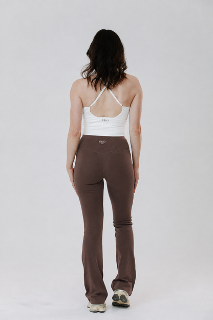 A tall woman viewed from behind wearing brown cotton high-waisted flared leggings and a white racerback tank top, standing against a light gray studio background.