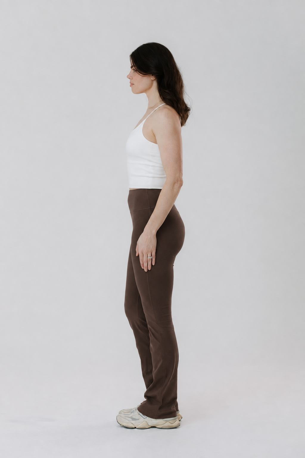 A tall woman in side profile wearing brown cottom high-waisted flared activewear pants, and a white cotton tank top, and chunky white sneakers, standing against a light grey studio background.