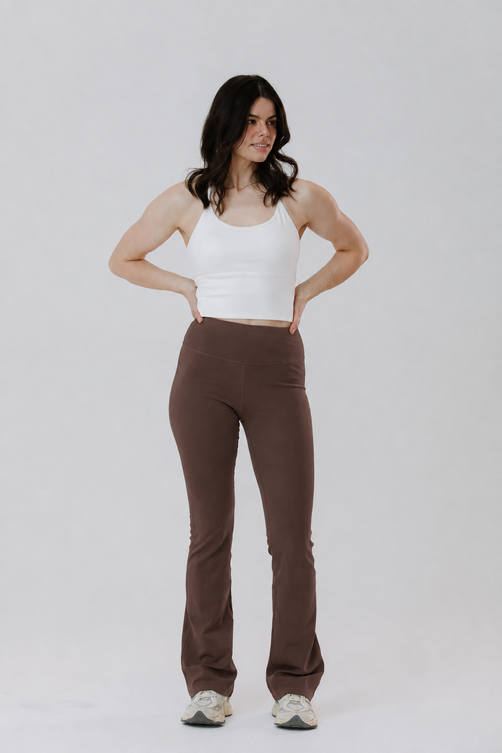 A tall woman modelling a pair of high-waisted, cotton brown flares activewear pants with a white tank top, standing against a light grey studio background.