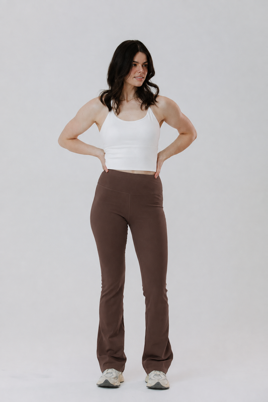 A tall woman modelling a pair of high-waisted, cotton brown flares activewear pants with a white tank top, standing against a light grey studio background.