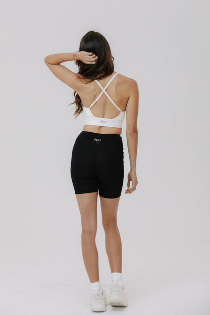 A woman is wearing a white cotton sports bra with crisscross straps and black cotton bike shorts, back view