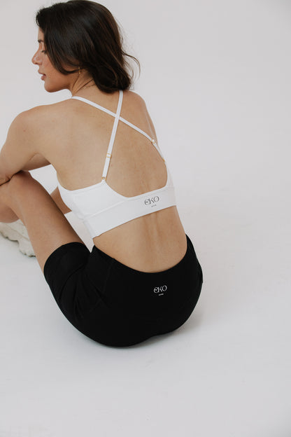 A woman seated shown from the back wearing a cotton white sports bra with thin crisscross straps and black cotton bike shorts.