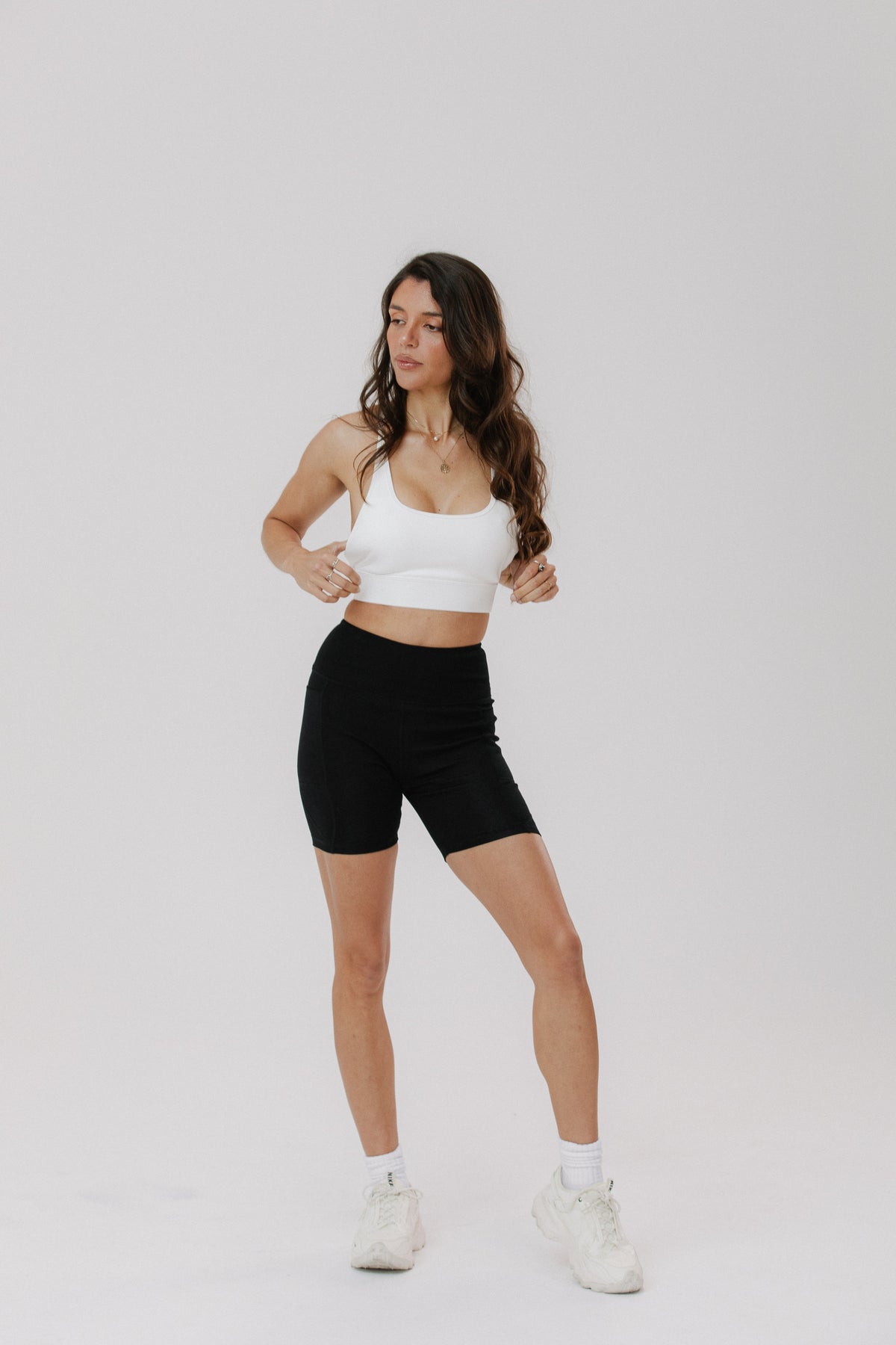 A woman modeling a white sports bra and black biker shorts against a plain light gray studio background.