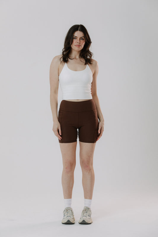 A woman stands against a light grey studio background wearing dark brown cotton fitted bike shorts, a white ribbed cotton tank top, white socks, and light-coloured sneakers.