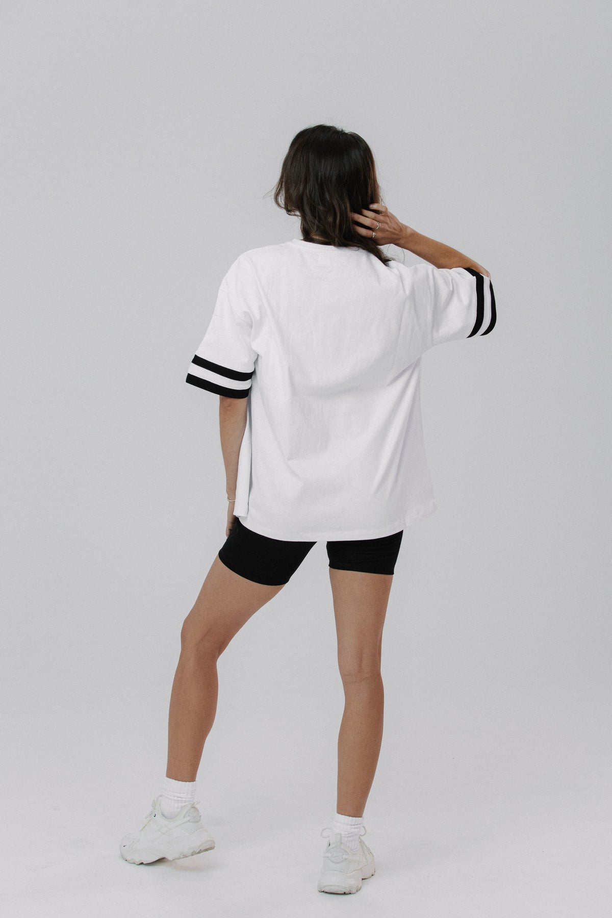 A woman is shown from behind wearing an oversized white t-shirt with black stripes on the sleeves, black fitted shorts, white socks, and white sneakers, set against a plain light gray studio background.