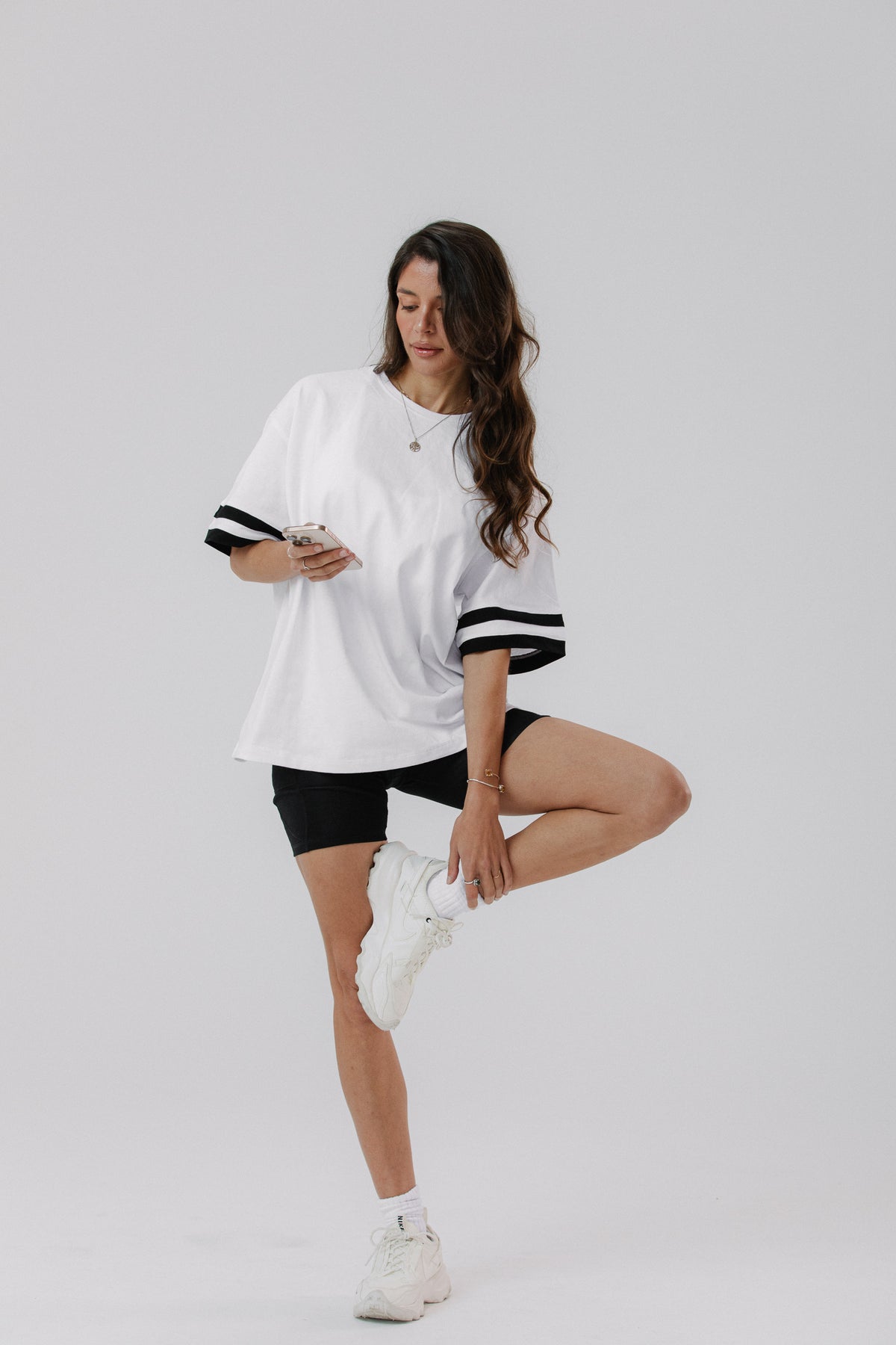 A woman posing in a white oversized t-shirt with black striped sleeve cuffs, black bike shorts, and white sneakers against a light gray studio background.