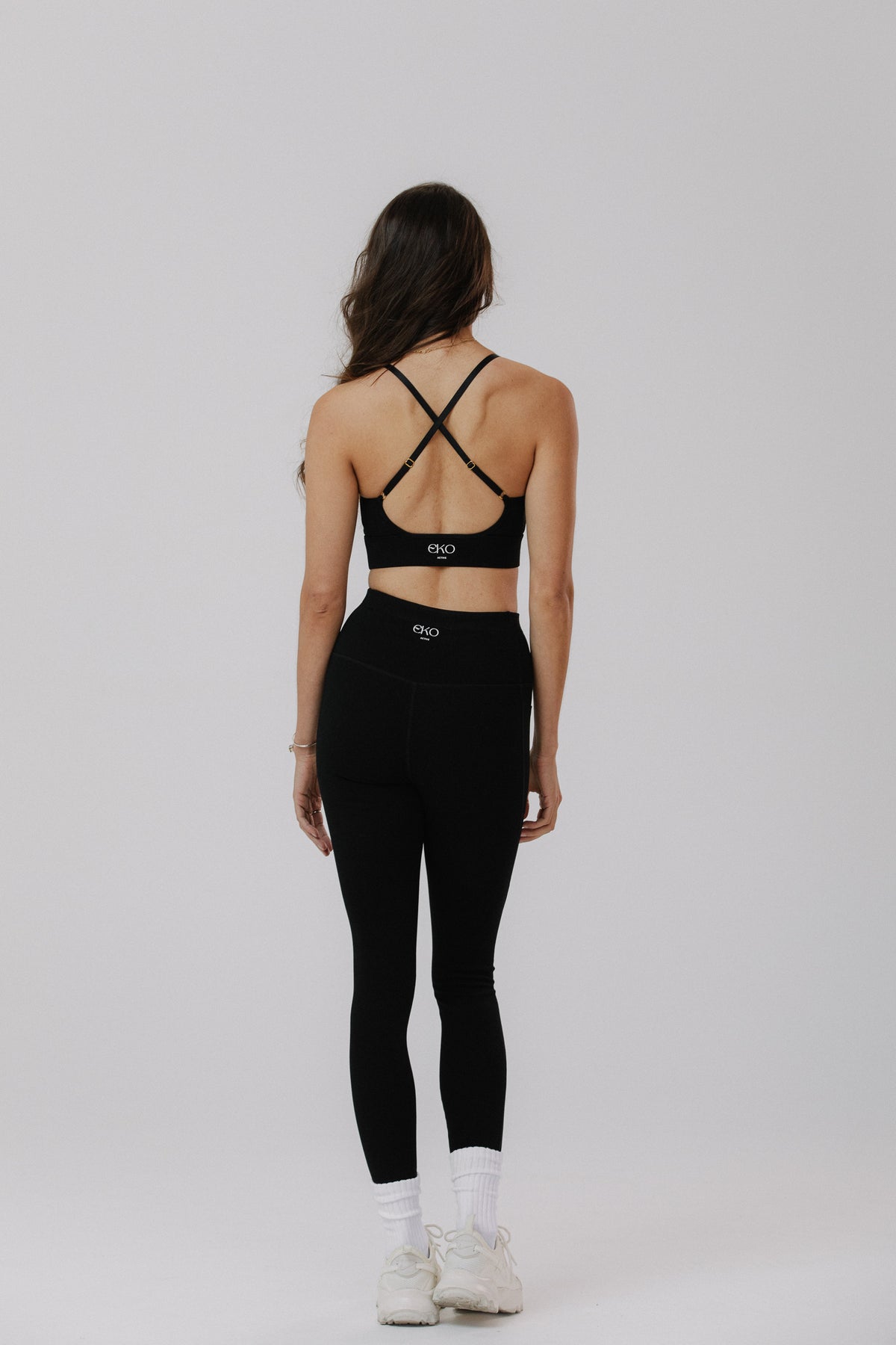 A woman is shown from the back wearing a black cotton sports bra with crisscross straps and matching black high-waisted cotton leggings, paired with white socks and white sneakers.