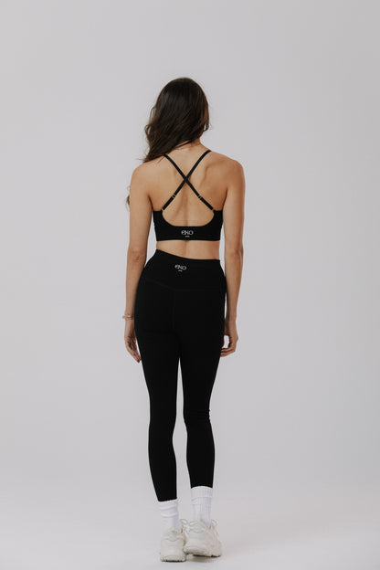 A woman is shown from the back wearing a black cotton sports bra with crisscross straps and matching black high-waisted cotton leggings, paired with white socks and white sneakers.