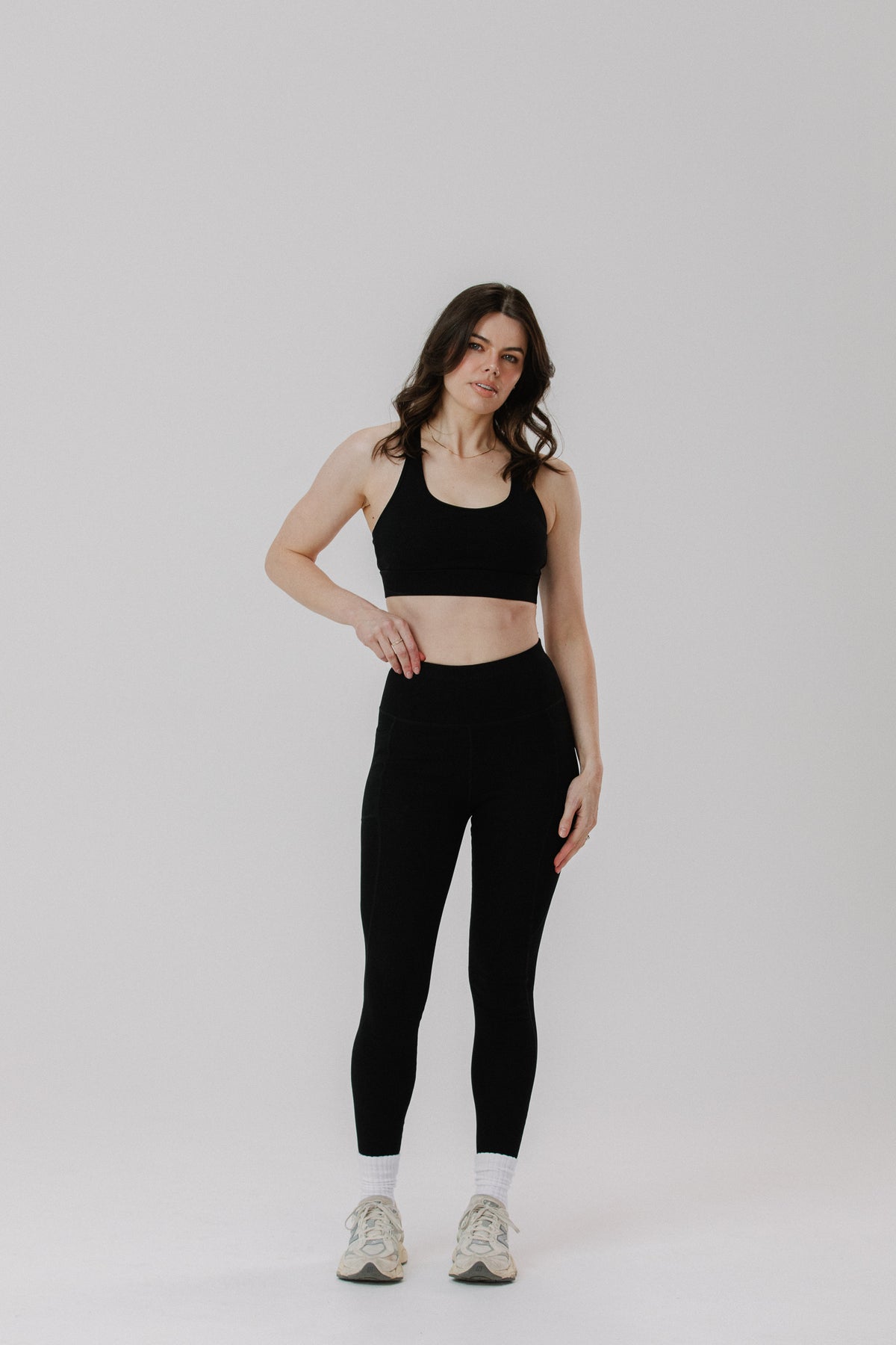 A tall woman modelling a black cotton sports bra and matching black cotton high-waisted leggings with white socks and light-coloured sneakers, standing against a plain light grey studio background.