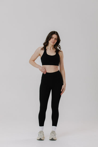 A tall woman modelling a black cotton sports bra and matching black cotton high-waisted leggings with white socks and light-coloured sneakers, standing against a plain light grey studio background.