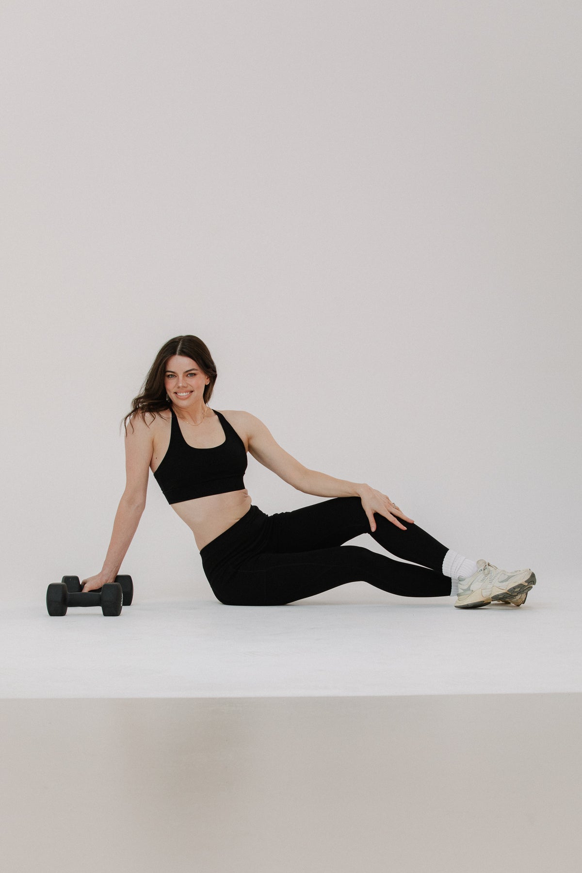 A tall woman in a black cotton sports bra and black cotton leggings sits on a white studio floor, holding a small black dumbbell in one hand and wearing white sneakers. EKO Active brand