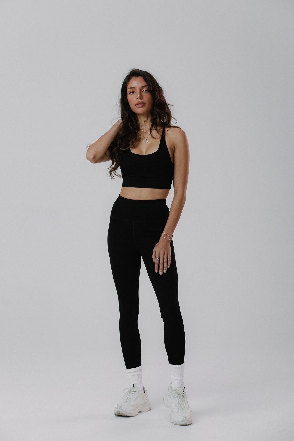 A woman modeling a black cotton activewear set consisting of a scoop-neck tank top and high-waisted black cotton leggings, paired with white socks and white sneakers.