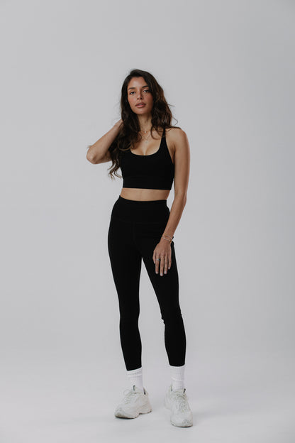 A woman modeling a black cotton activewear set consisting of a scoop-neck tank top and high-waisted black cotton leggings, paired with white socks and white sneakers.