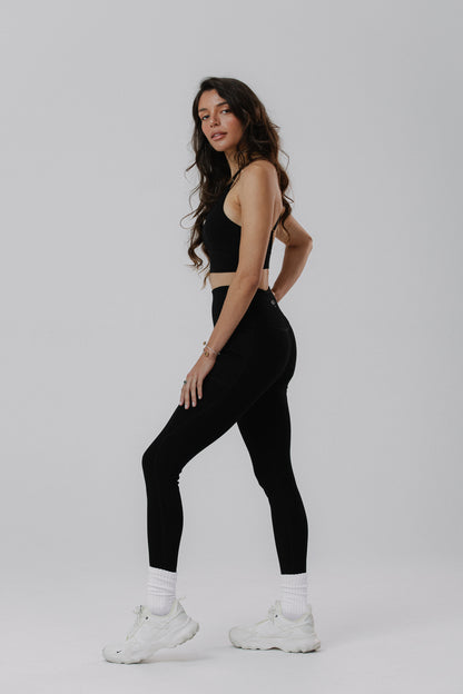A woman modeling black cotton activewear set, consisting of a black cotton sports bra and black cotton high-waisted leggings, paired with white socks and white chunky sneakers