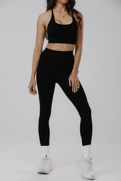 A woman modeling a black sports bra and matching high-waisted black leggings with white socks and white sneakers, photographed against a light gray studio background.