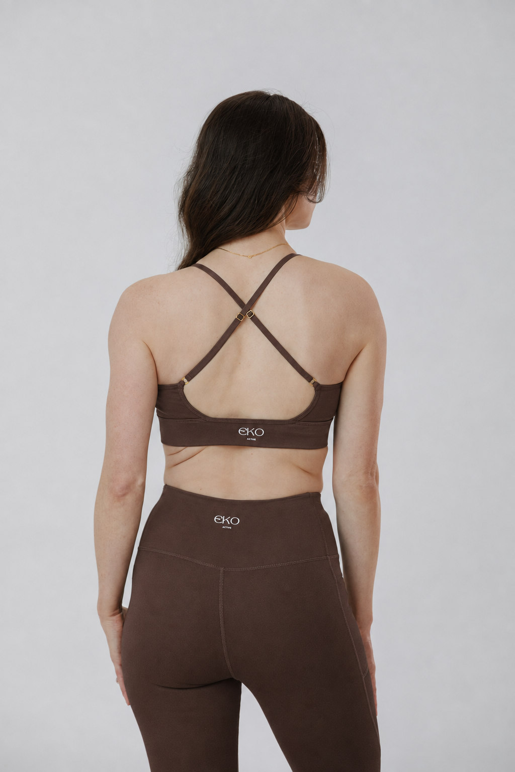 A woman shown from the back wearing a brown sports bra with crisscross straps and matching high-waisted leggings against a light gray studio background.