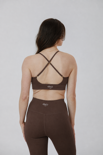 A woman shown from the back wearing a brown sports bra with crisscross straps and matching high-waisted leggings against a light gray studio background.