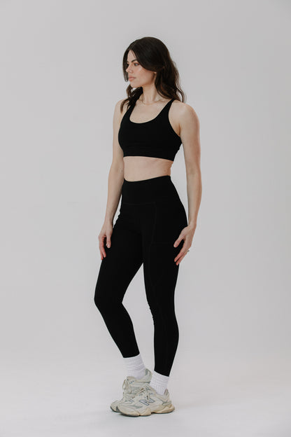 A tall woman modeling a black sports bra and matching black high-waisted cotton leggings with white socks and white sneakers, standing against a light grey studio background.