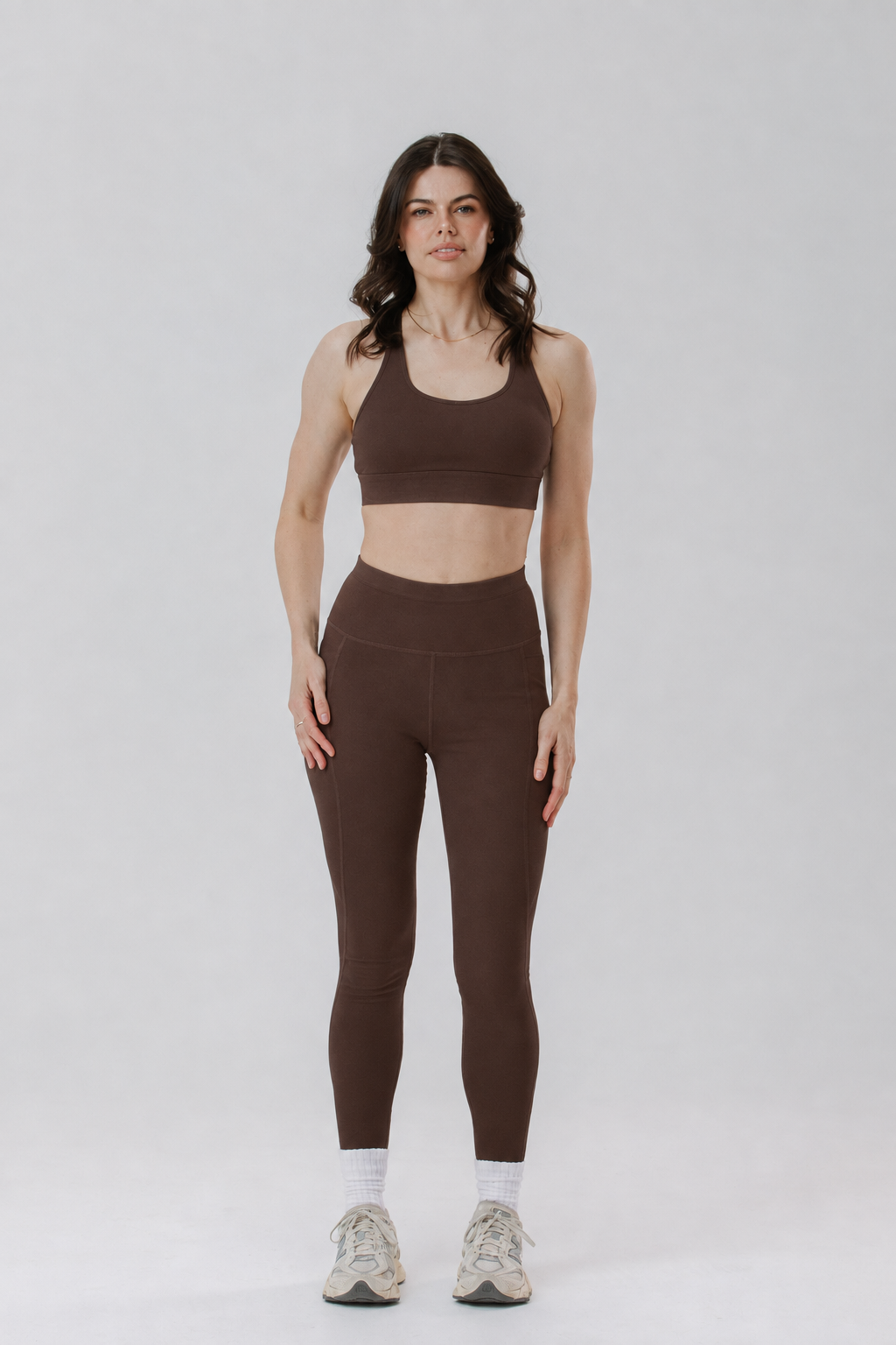 A tall woman modelling a matching brown activewear set consisting of a sports bra and tall length high-waisted cotton leggings, paired with white socks and white sneakers, against a light grey studio background.