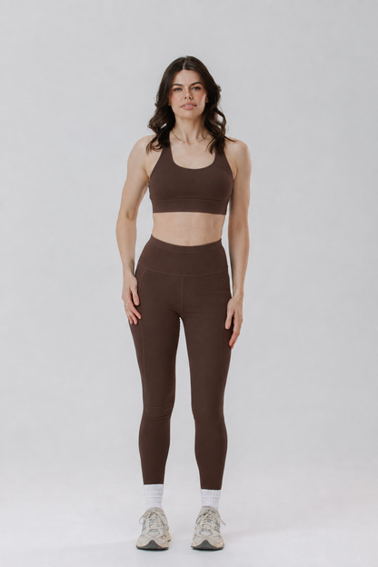 A tall woman modelling a matching brown activewear set consisting of a sports bra and tall length high-waisted cotton leggings, paired with white socks and white sneakers, against a light grey studio background.
