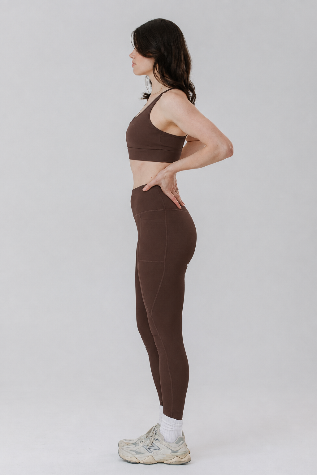 A tall woman in side profile wearing a matching brown cotton activewear set consisting of a sports bra and high-waisted cotton leggings in tall length, paired with white socks and white sneakers, against a light grey studio background.