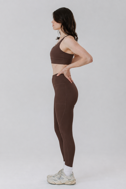 A tall woman in side profile wearing a matching brown cotton activewear set consisting of a sports bra and high-waisted cotton leggings in tall length, paired with white socks and white sneakers, against a light grey studio background.