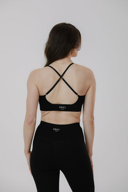 A woman shown from the back wearing a black cotton sports bra with crisscross straps and black leggings, photographed against a plain light gray studio background.