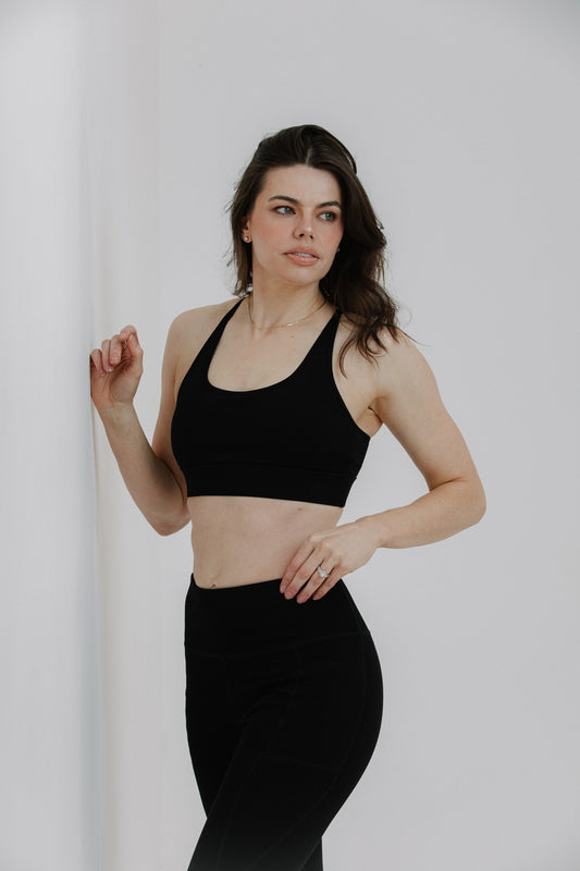 A woman modeling a black cotton sports bra with built-in cups and matching black leggings against a plain light grey studio background.