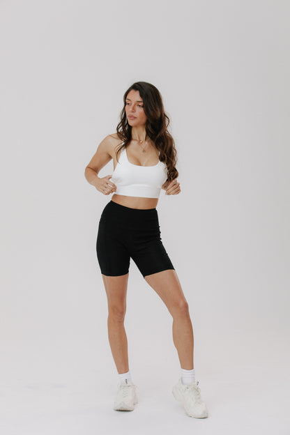 A woman modeling a white sports bra and black biker shorts against a plain light gray studio background.