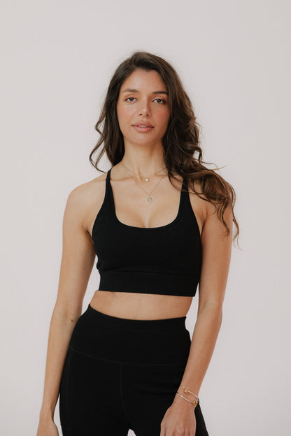 A woman modelling a black cotton sports bra and matching black high-waisted leggings against a plain light grey studio background.