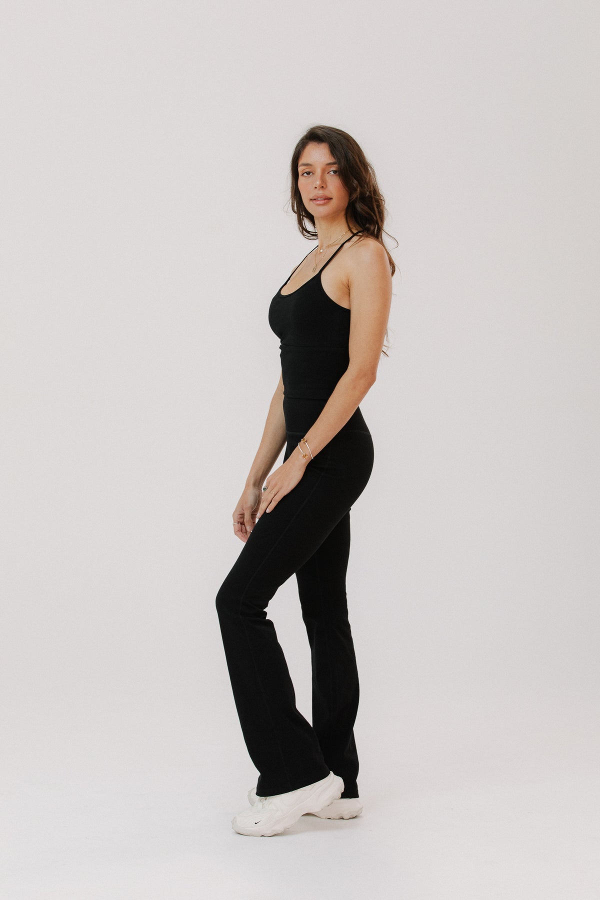 A woman modeling a black cotton tank top and matching black flared pants, paired with white sneakers, standing against a seamless white studio background.