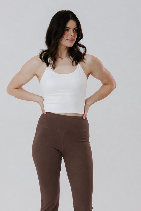 A woman modelling a pair of high-waisted, straight-leg brown pants with a white cotton tank top, standing against a light grey studio background.